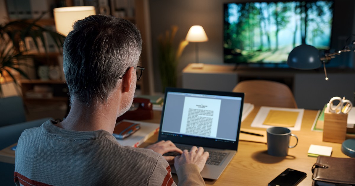 Man with short gray hair and glasses sitting at a desk in a warmly lit home office, typing on a laptop displaying a document, with a mug, phone, papers, and desk lamp nearby, while a television in the background shows a peaceful forest scene, suggesting focused writing or working in the evening.
