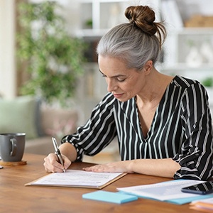 Woman sitting at a table, focused on writing notes on paper with a pen, with notebooks, sticky notes, and a mug nearby. She is planning, and organizing ideas in a calm home setting.